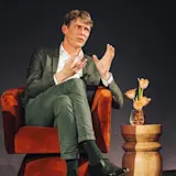 Foto van Huub van der Meulen terwijl hij spreekt voor een publiek. Hij zit op een stoel op een podium.