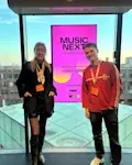 Twee personen staan voor een kleurrijk scherm tijdens het Music Next-evenement in een moderne evenementenlocatie.