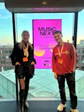 Twee personen staan voor een kleurrijk scherm tijdens het Music Next-evenement in een moderne evenementenlocatie.
