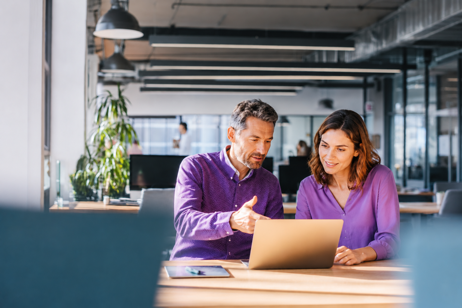 Man in paars overhemd legt iets uit op een laptop aan een vrouwelijke collega in een paarse blouse, aan een houten tafel in een modern kantoor.