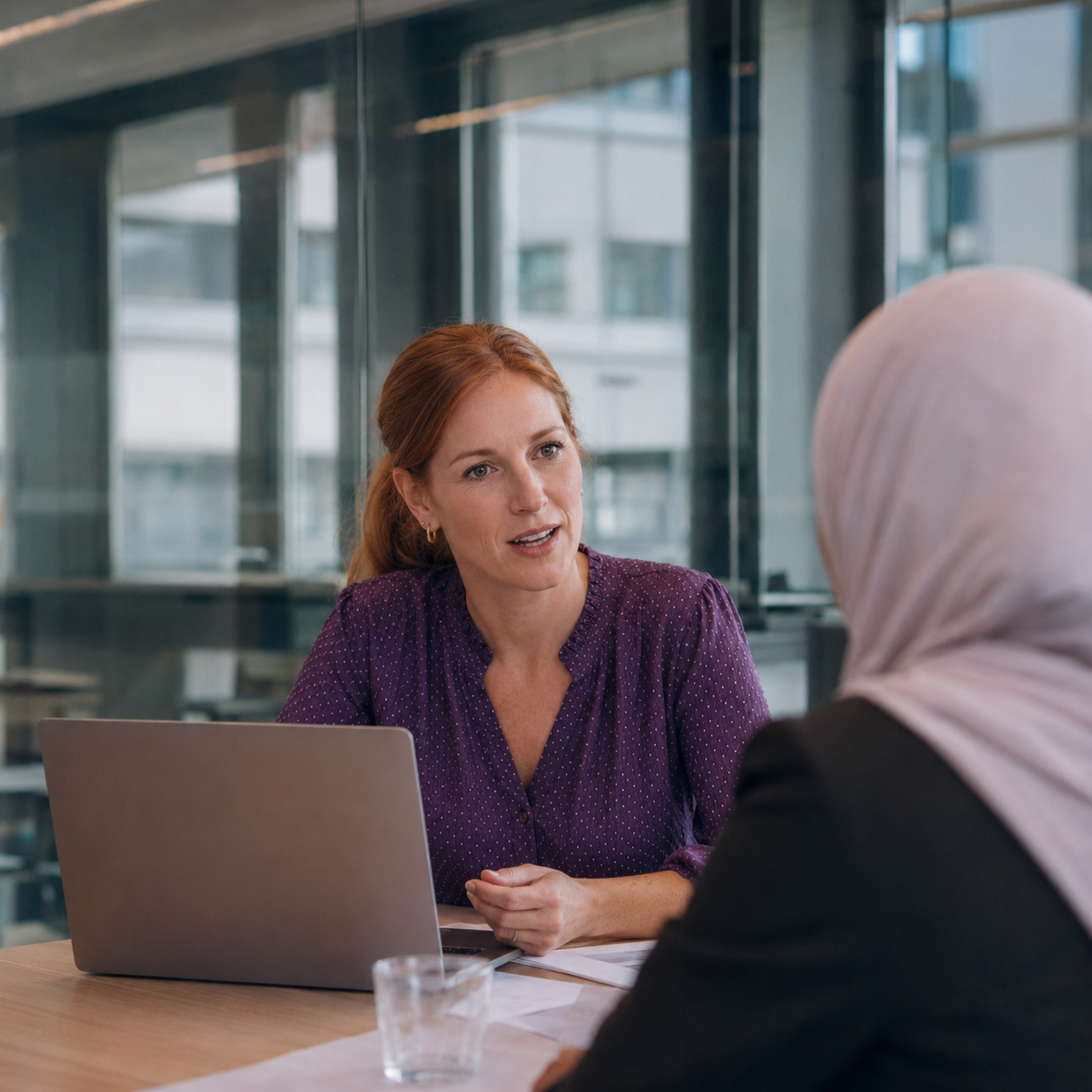 wee collega's voeren een gesprek aan tafel, waarbij een vrouw in een paarse blouse tegenover een collega met hoofddoek zit.