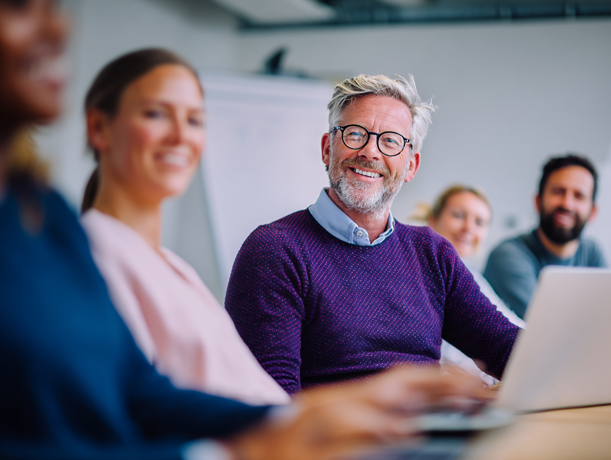 Collega's nemen deel aan een overleg in een moderne vergaderruimte. Focus is op een man met wit haar, paarse trui en een ronde bril.