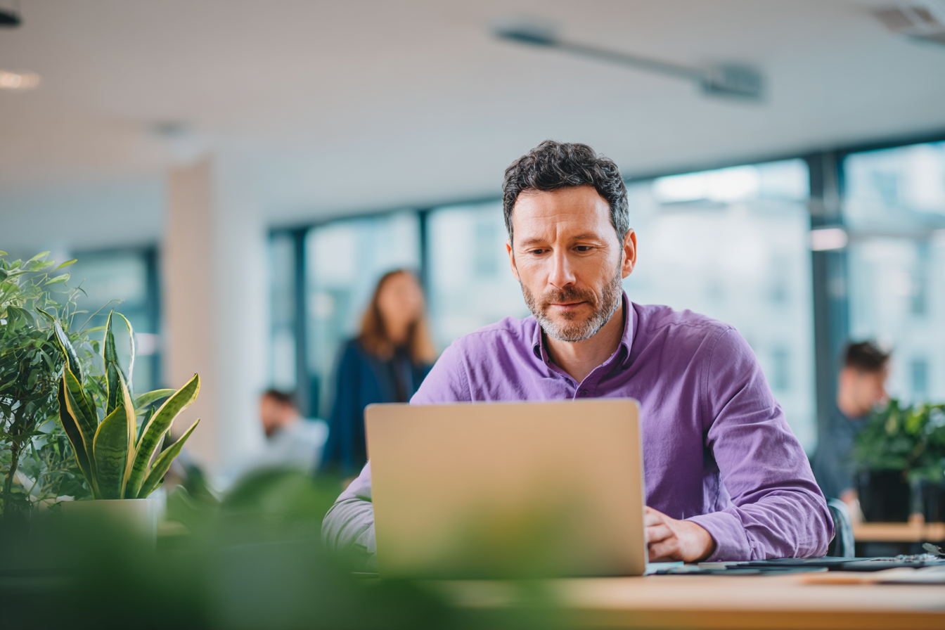 Man in een donkerpaarse blouse werkt achter een laptop aan een bureau in een licht en modern kantoor. Op de voorgrond zijn planten zichtbaar en op de achtergrond lopen collega’s door de kantoorruimte.