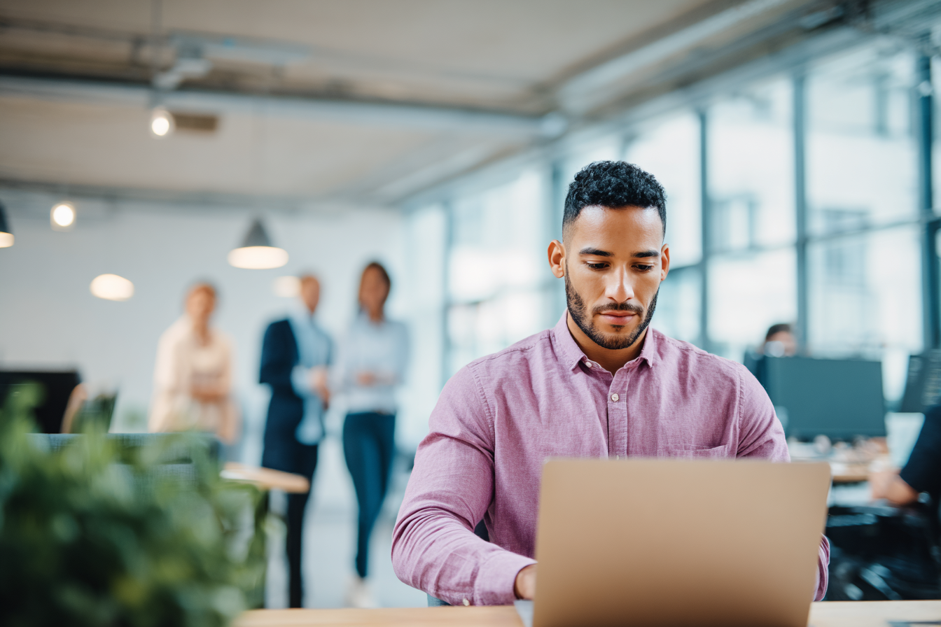 Man in een donkerpaarse blouse werkt achter een laptop aan een bureau in een licht en modern kantoor. Op de voorgrond zijn planten zichtbaar en op de achtergrond lopen collega’s door de kantoorruimte.