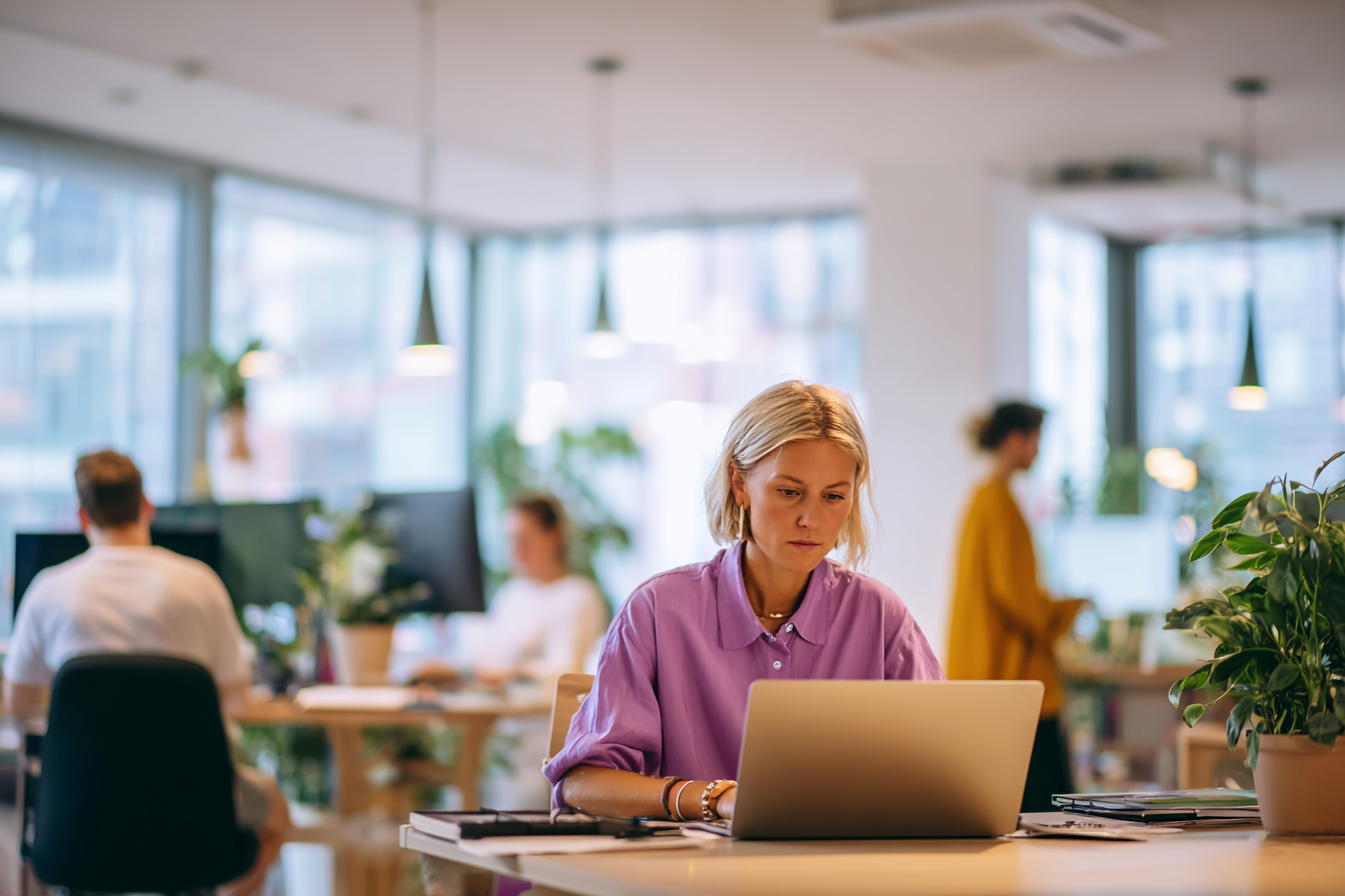 Vrouw in een donkerpaarse blouse werkt achter een laptop aan een bureau in een licht en modern kantoor. Op de voorgrond zijn planten zichtbaar en op de achtergrond lopen collega’s door de kantoorruimte.