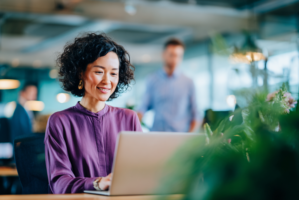 Vrouw een donkerpaarse blouse werkt achter een laptop aan een bureau in een licht en modern kantoor. Op de voorgrond zijn planten zichtbaar en op de achtergrond lopen collega’s door de kantoorruimte.