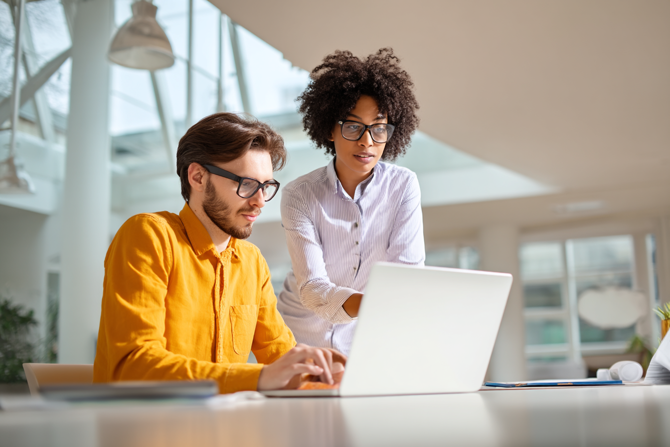 Man in gele blouse en vrouw met krullend haar en bril kijken samen naar een laptopscherm.