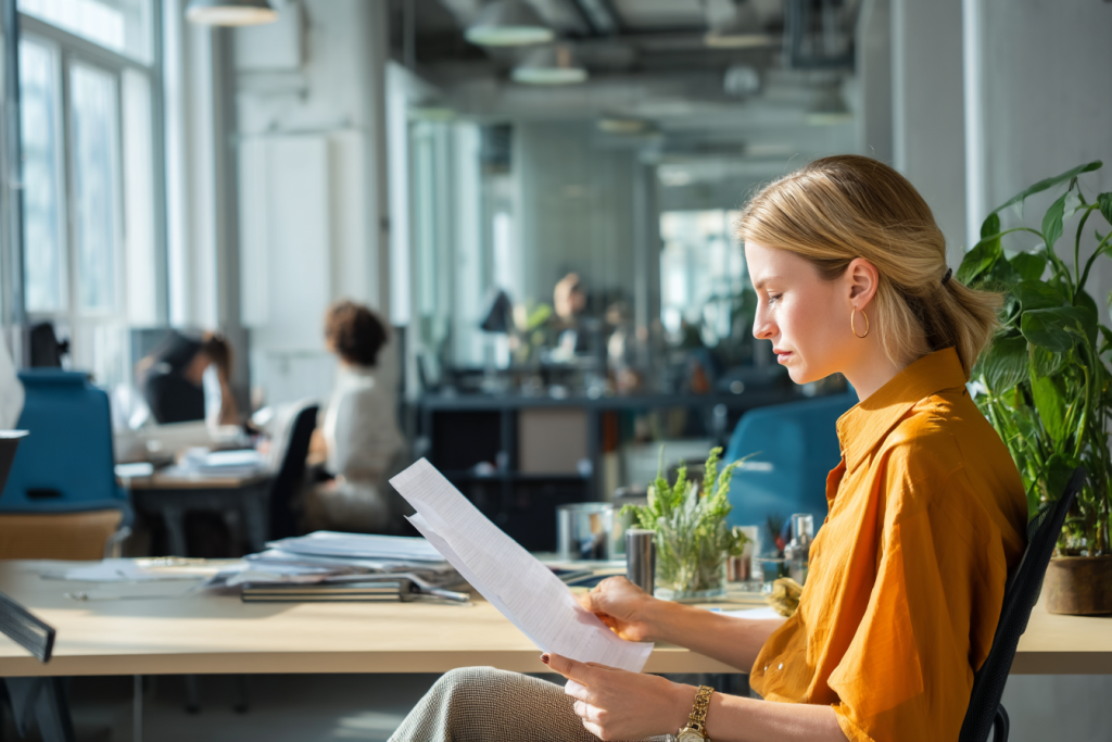 Vrouw met blond haar in oranje blouse leest zittend een document in een kantoor met veel glas en planten.
