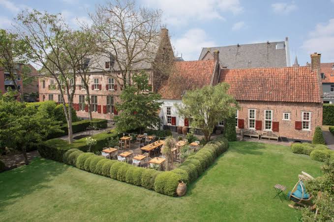 Historisch bakstenen gebouw met rode luiken, omringd door een grote groene tuin met hagen en een terras met houten tafels.