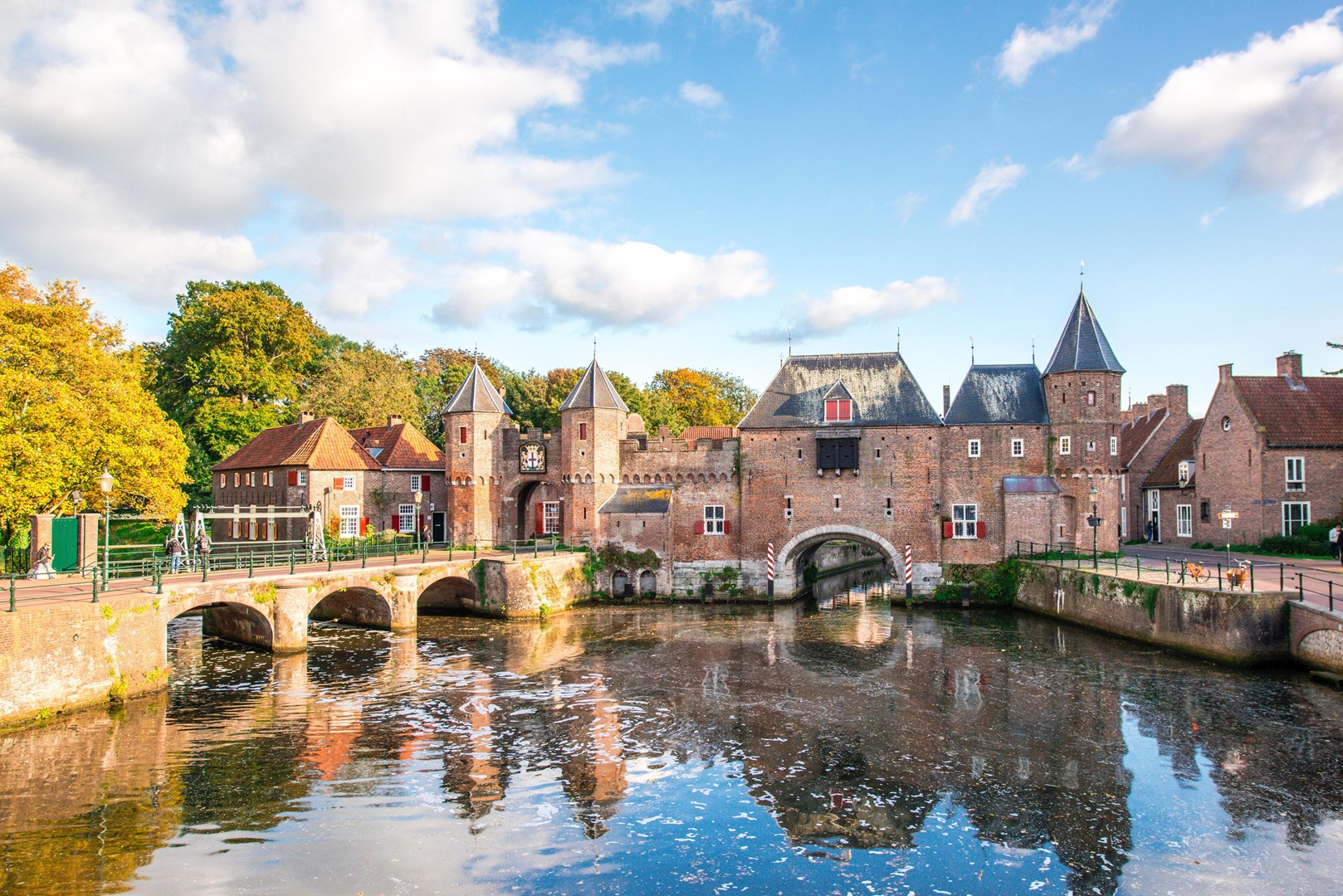 Historische Koppelpoort in Amersfoort met bakstenen torens en stadsmuren, weerspiegeld in het water onder een blauwe lucht.